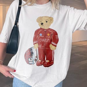 以下が含まれることがあります： 白い半袖Tシャツ。赤いレーシングスーツを着たベージュのテディベアのグラフィックが描かれており、スーツには「CHARLES」の文字と「16」の数字がある。クマの横には、側面に「16」の数字が見えるレーシングヘルメットが配置されている。