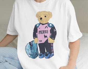 Może przedstawiać: Biały t-shirt z grafiką beżowego pluszowego misia ubranego w różowo-czarny kombinezon wyścigowy z napisem PIERRE, małym czarnym logo i numerem 10. Miś trzyma niebieski kask wyścigowy i ma na sobie niebieskie buty wyścigowe.