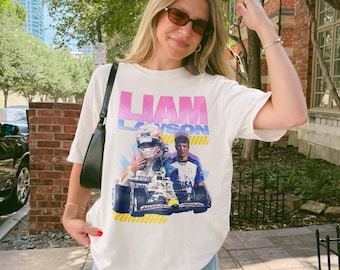 Może przedstawiać: Biały t-shirt z krótkim rękawem z nadrukiem graficznym przedstawiającym bolid Formuły 1, kierowcę w kasku oraz portret Liama Lawsona. Napis LIAM LAWSON jest wydrukowany pogrubionymi różowymi i niebieskimi literami nad grafiką wyścigową.