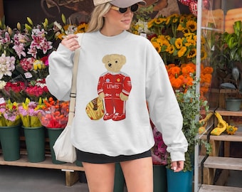 Puede incluir: Una sudadera blanca de manga larga con el gráfico de un oso de peluche vestido con un traje de carreras rojo con el nombre LEWIS impreso en el pecho, sosteniendo un casco amarillo. La sudadera tiene cuello redondo y puños y dobladillo acanalados.