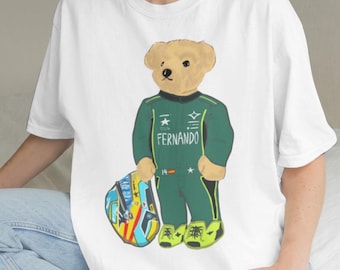 Może przedstawiać: Biały t-shirt z grafiką jasnobrązowego pluszowego misia ubranego w ciemnozielony kombinezon wyścigowy z nadrukowanym na piersi imieniem FERNANDO. Miś trzyma w jednej łapie kolorowy kask wyścigowy z niebieskimi, żółtymi i czerwonymi akcentami.