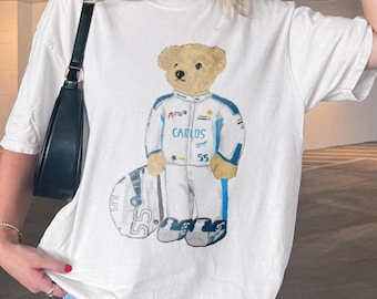 Może przedstawiać: Biały t-shirt z krótkim rękawem z grafiką beżowego misia ubranego w biało-niebieski kombinezon wyścigowy z nadrukowanym na piersi imieniem CARLOS i numerem 55. Miś stoi obok białego kasku wyścigowego z widocznym numerem 55.
