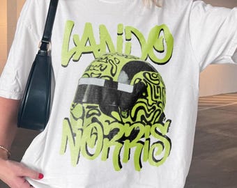 Może przedstawiać: Biały t-shirt z krótkim rękawem z nadrukiem graficznym kasku wyścigowego w limonkowo-czarny abstrakcyjny wzór. Napis "LANDO" znajduje się nad kaskiem, a "NORRIS" pod nim, wykonany limonkowymi literami w stylu graffiti.