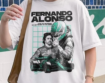 Może przedstawiać: Biały t-shirt z nadrukiem graficznym Fernando Alonso. Projekt zawiera portret kierowcy, postać w kasku wyścigowym, bolid Formuły 1 oraz tekst "FERNANDO ALONSO" i "WHEN YOU ARE SECOND, YOU ARE THE FIRST OF THE LOSERS."