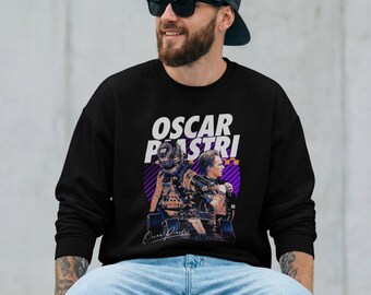 May include: A black long-sleeve sweatshirt featuring a graphic print of Formula 1 driver Oscar Piastri. The design includes the text OSCAR PIASTRI in white, a depiction of the driver in a racing suit and helmet, and a race car on a purple background.