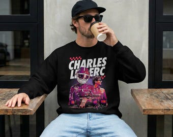 Może przedstawiać: Czarna bluza z długim rękawem z nadrukiem graficznym przedstawiającym Charlesa Leclerca. Wzór zawiera biały napis "CHARLES LECLERC", wizerunek kierowcy w kasku wyścigowym, samochód wyścigowy oraz portret w odcieniach czerwieni i fioletu.
