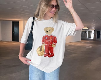 Może przedstawiać: Biały t-shirt z krótkim rękawem z grafiką misia ubranego w czerwony kombinezon wyścigowy z napisem LEWIS na piersi. Miś trzyma żółto-brązowy kask wyścigowy. Koszulka jest zestawiona z jasnoniebieskimi dżinsami.