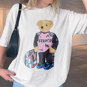 以下が含まれることがあります： 白の半袖Tシャツ。ベージュのテディベアが描かれており、黒とピンクのレーシングスーツを着用しています。スーツには「FRANCO」、数字の「43」、小さな雄牛のロゴがプリントされています。クマの横には、カラフルな模様の円形の物体が配置されています。
