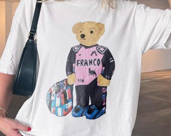 Może przedstawiać: Biały t-shirt z krótkim rękawem z grafiką beżowego misia ubranego w czarno-różowy kombinezon wyścigowy z napisem FRANCO, numerem 43 i małym logo byka. Miś stoi obok kolorowego, wzorzystego obiektu o okrągłym kształcie.