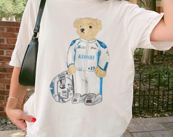 Może przedstawiać: Biały t-shirt z krótkim rękawem z grafiką jasnobrązowego misia ubranego w biało-niebieski kombinezon wyścigowy z napisem "ALEXANDER V2 *23" na piersi, stojącego obok pasującego biało-niebieskiego kasku wyścigowego.