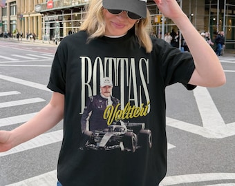 Może przedstawiać: Czarny t-shirt z krótkim rękawem z nadrukiem graficznym przedstawiającym kierowcę wyścigowego i bolid Formuły 1. Napis BOTTAS jest wydrukowany dużymi białymi literami nad kierowcą, a imię Valtteri żółtą kursywą pod zdjęciem kierowcy.