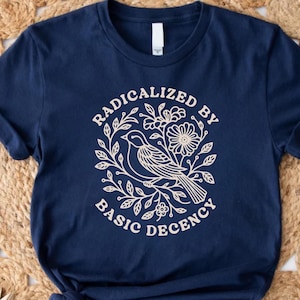 May include: Navy blue t-shirt featuring a cream-colored graphic of a bird, flowers, and leaves, with the text "RADICALIZED BY BASIC DECENCY" in a circular design. The shirt is made of soft material.