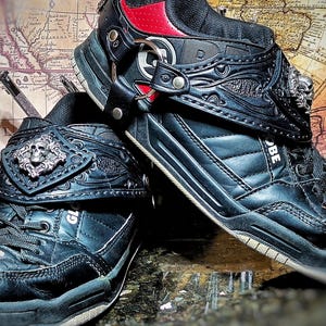 Puede incluir: Par de zapatillas de skate Globe negras con correas decorativas con detalles de calaveras. Las zapatillas tienen un aspecto de cuero con detalles rojos y plateados. El fondo incluye un mapa vintage.