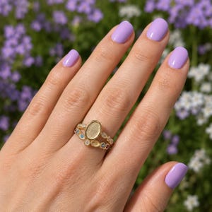 Può includere: Primo piano di una mano che indossa due anelli d'oro. L'anello superiore ha un'incastonatura ovale, mentre l'anello inferiore presenta piccole pietre preziose colorate. Lo sfondo è sfocato, mostrando fiori viola e bianchi.