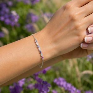 May include: A delicate silver bracelet adorned with alternating gemstones of blue, white, and red. The bracelet is displayed on a wrist against a blurred background of purple flowers and greenery.