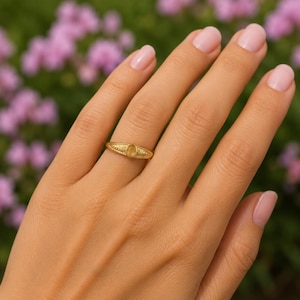 May include: A gold ring with an oval-shaped, light-colored stone. The ring has a delicate design on the band and is set against a blurred background of pink flowers.