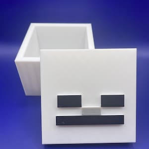 May include: A white, square box with a lid featuring a stylized skeleton face design. The lid has black and gray rectangular shapes for the eyes and mouth. The box is open, revealing its interior.