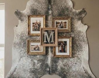 May include: A large gray and white speckled cowhide rug mounted on a wall as a backdrop for four ornate gold picture frames containing family photographs and a central square wooden frame featuring a large, metallic letter M in the center.