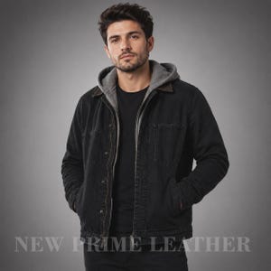 Op de afbeelding: Een zwart spijkerjack met een grijze capuchon, een klassieke kraag en een knoopsluiting aan de voorzijde. Het jack heeft twee borstzakken en lange mouwen. De tekst NEW PRIME LEATHER staat in het wit onderaan de afbeelding gedrukt.