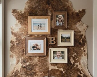May include: A large brown and white cowhide rug mounted on a wall as a backdrop for a gallery wall arrangement. The display features four rectangular picture frames in various wood tones and cream colors, along with a wooden letter B centerpiece.