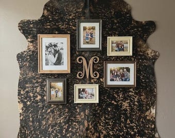 May include: A large, irregular, brown and black mottled cowhide rug mounted on a wall, featuring five rectangular picture frames of varying sizes and colors, and a central wooden monogram letter M with decorative scrollwork accents.