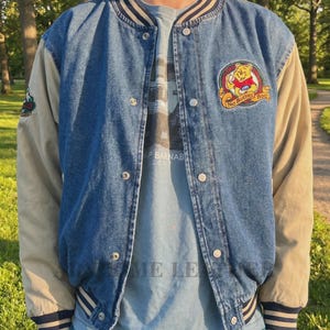 Puede incluir: Una chaqueta universitaria de mezclilla azul con mangas largas beige, cuello y puños de canalé a rayas y botones a presión plateados. El pecho izquierdo presenta un parche bordado de Winnie the Pooh con el texto No Bother Bear.