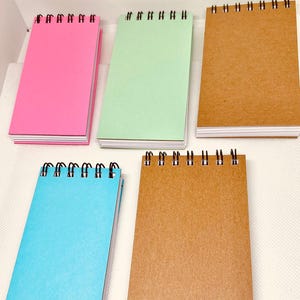 Puede incluir: Cinco pequeños blocs de notas con espiral en varios colores: rosa, verde, marrón, azul y otro marrón. Los blocs de notas son rectangulares con un borde blanco y encuadernación en espiral negra en la parte superior. El papel parece en blanco.