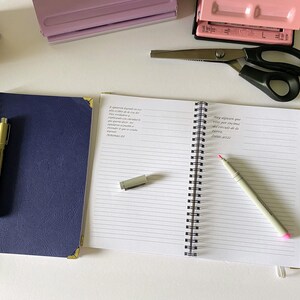 May include: An overhead shot of stationery items including a navy blue spiral notebook with a gold pen, an open notebook with text, scissors, a ruler, and a pink hole punch. A rainbow pencil and a small decorative box are also visible.