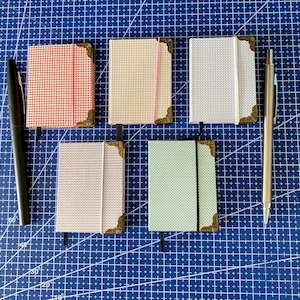 Puede incluir: Cinco cuadernos pequeños en varios colores, incluyendo rojo, beige, blanco, rosa y verde, están dispuestos sobre una superficie de cuadrícula azul. Cada cuaderno tiene detalles decorativos en las esquinas y un cierre de banda elástica. También hay dos bolígrafos.