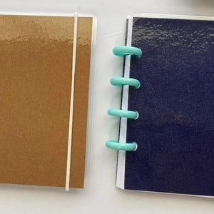 May include: Two small notebooks with colorful disc binding. One has a brown cover and a white elastic band, the other has a dark blue cover and a black elastic band. A pink ruler is on the left.