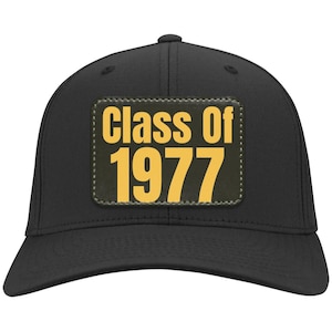 Może przedstawiać: Czarna czapka z daszkiem z zakrzywionym daszkiem. Czapka posiada prostokątną naszywkę z żółtym napisem "Class Of 1977", obramowanym ciemniejszym odcieniem, na zielonym tle. Wykonana z wytrzymałej tkaniny.