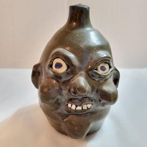 May include: A ceramic jug shaped like a grotesque face. The jug is a dark olive green color with bulging cheeks, a bulbous nose, and a small mouth with visible teeth. The eyes are white with blue pupils.