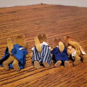 Puede incluir: Cuatro figuras de animales de juguete marrones, cada una con un atuendo diferente. Una figura lleva un abrigo azul, otra un pantal&oacute;n a rayas y una tercera un traje azul. La cuarta figura lleva una camisa color melocot&oacute;n.