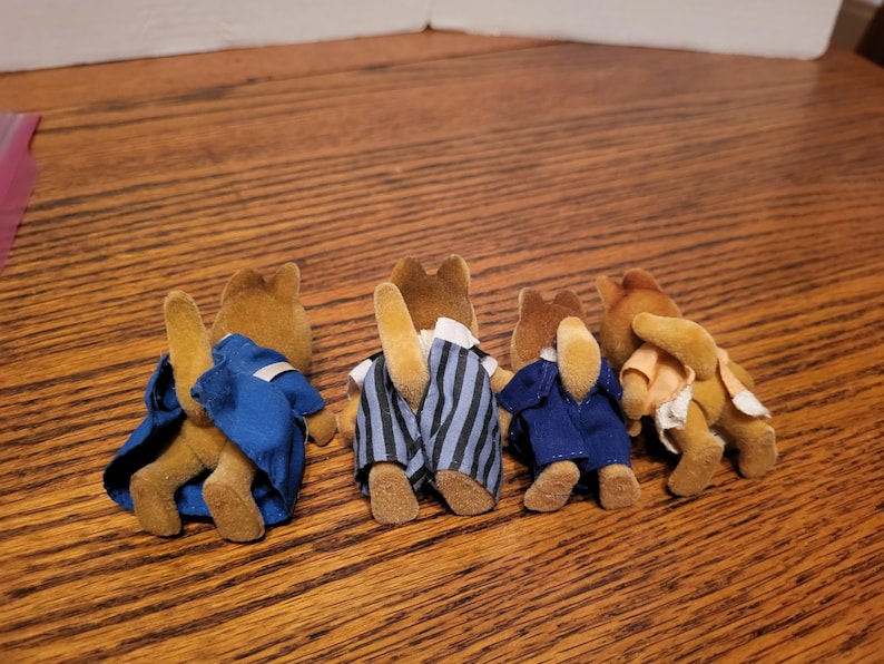 Puede incluir: Cuatro figuras de animales de juguete marrones, cada una con un atuendo diferente. Una figura lleva un abrigo azul, otra un pantal&oacute;n a rayas y una tercera un traje azul. La cuarta figura lleva una camisa color melocot&oacute;n.