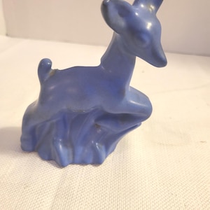 May include: A blue ceramic deer figurine. The deer is in a seated position with its head and ears upright. The figurine has a smooth, glossy finish and is a solid shade of blue. The deer is resting on a base that resembles a stylized cloud.