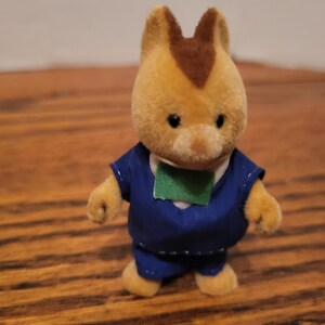 Puede incluir: Un peque&ntilde;o juguete de color marr&oacute;n claro con una mancha marr&oacute;n en la cabeza, que lleva un atuendo azul con un parche verde. El juguete tiene una expresi&oacute;n amigable y est&aacute; de pie sobre una superficie de madera.