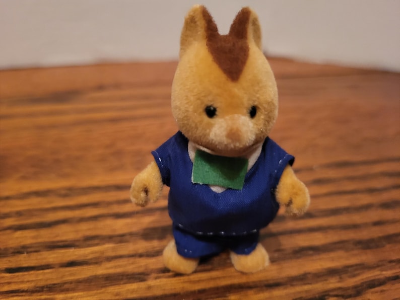 Puede incluir: Un peque&ntilde;o juguete de color marr&oacute;n claro con una mancha marr&oacute;n en la cabeza, que lleva un atuendo azul con un parche verde. El juguete tiene una expresi&oacute;n amigable y est&aacute; de pie sobre una superficie de madera.