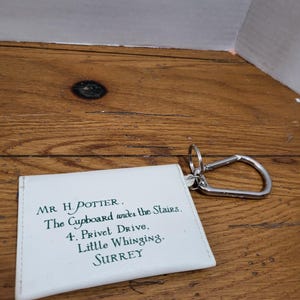 May include: A white rectangular keychain featuring dark green printed text that reads: MR H. POTTER. The Cupboard under the Stairs. 4. Privet Drive, Little Whinging, SURREY. The item includes a silver-colored metal carabiner and a circular ring.