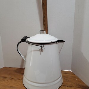 May include: A white enamel coffee pot with a black rim, a black handle with a wooden grip, and a matching lid. A vertical wooden ruler shows the height is approximately 12 inches, and a horizontal ruler shows the base is 8 inches wide.