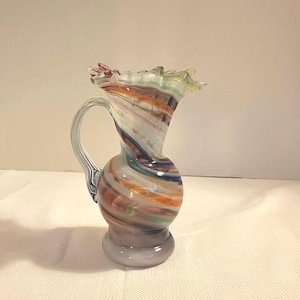 Może przedstawiać: Mały, dekoracyjny szklany dzbanek z falistym brzegiem i uchwytem. Dzbanek ma wirujący wzór w kolorach pomarańczowym, niebieskim, białym i brązowym. Podstawa jest w kolorze jasnofioletowym.
