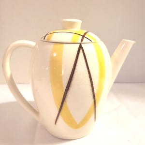 Puede incluir: Tetera de cerámica color crema con tapa, con diseños geométricos amarillos y marrones. La tetera tiene un asa curva y un pico. La tapa tiene un pequeño pomo. Un artículo de cocina vintage.