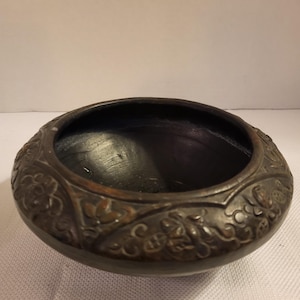 Puede incluir: Un cuenco de color marrón oscuro, de estilo antiguo, con un diseño floral en relieve alrededor del borde. El cuenco tiene una forma ancha y poco profunda y un interior liso y oscuro. El exterior tiene un brillo metálico.