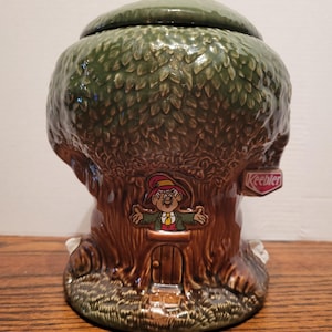 May include: A ceramic cookie jar shaped like a tree, with a green leafy top and a brown trunk. A cartoon elf figure is on the front, with a door and the word "Keebler" on a sign. The jar is on a wooden surface.