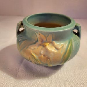 Puede incluir: Una pequeña maceta redonda de cerámica con un diseño floral en azul claro y dorado. La maceta tiene dos asas y una superficie texturizada. El diseño floral está en relieve, lo que añade dimensión a la pieza. El interior es de color marrón oscuro.