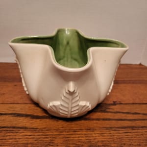 May include: A white ceramic pitcher with a green interior. The pitcher has a unique shape with a spout and a leaf design on the front. The pitcher is likely used for serving beverages or as a decorative item.