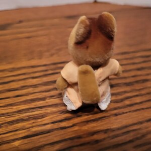 Puede incluir: Peque&ntilde;a figura de oso de juguete vintage con cabeza y orejas marrones, vestida con un atuendo de color beige claro. El oso est&aacute; posado con los brazos envueltos alrededor de s&iacute; mismo. El juguete est&aacute; sobre una superficie de madera.