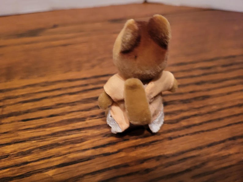 Puede incluir: Peque&ntilde;a figura de oso de juguete vintage con cabeza y orejas marrones, vestida con un atuendo de color beige claro. El oso est&aacute; posado con los brazos envueltos alrededor de s&iacute; mismo. El juguete est&aacute; sobre una superficie de madera.