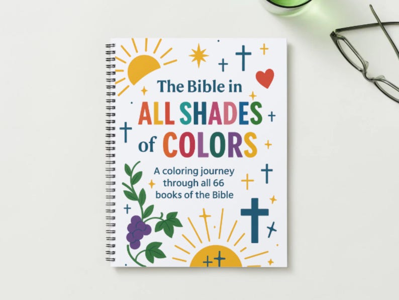 May include: A spiral-bound colouring book titled "The Bible in All Shades of Colors." The cover features colourful text, crosses, a heart, sunbursts, and illustrations of grapes and leaves. The book is designed for colouring through all 66 books of the Bible.
