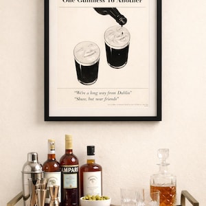 May include: A framed print with the text "One Guinness To Another" above two glasses of stout. Below, a bar cart holds bottles of liquor, a cocktail shaker, jiggers, a bowl of olives, and glassware. A decanter is also present.