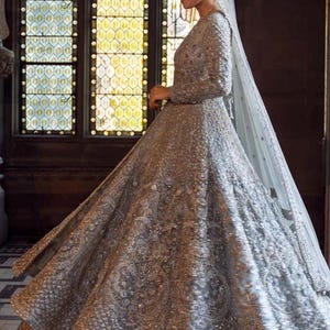 May include: A silver, long-sleeved wedding gown with intricate beading and embroidery. The dress features a full skirt and a long, flowing veil. The gown is worn by a person standing in front of a large window with stained glass.
